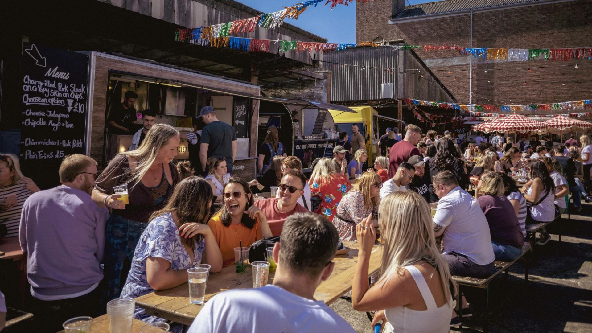 people socialising in picnic tables surrounded by food trucks